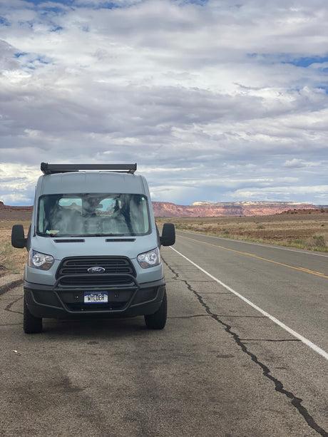 An RV rental, a grey Ford Transit camper van, is parked on a scenic desert road with distant red cliffs and a cloudy sky.