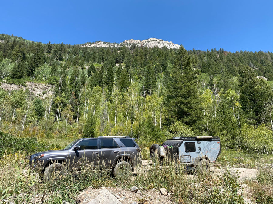 Overland RV rental adventure: Gray SUV towing a trailer on a dirt road through scenic mountain forests.
