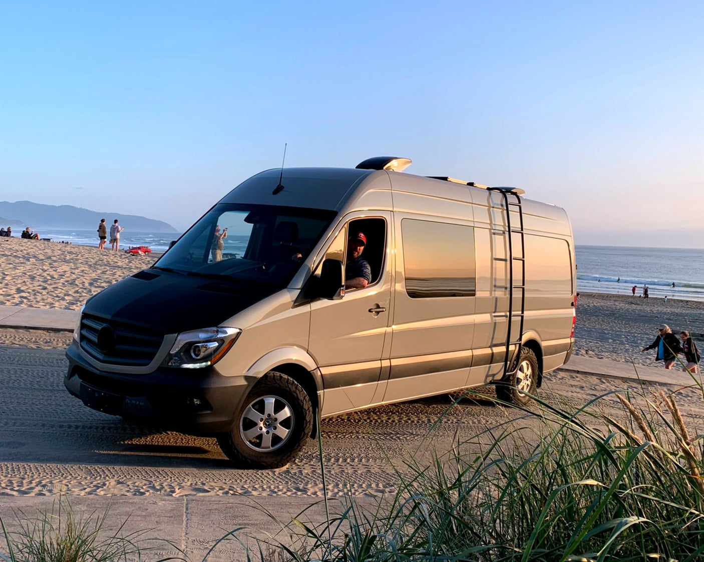Enjoy beach travel! RV rental: a light gray Mercedes Sprinter camper van on a sandy path next to the ocean at sunset.