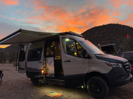 Gorgeous sunset over an RV rental camper van campsite. Awning extended, lights on, revealing a welcoming outdoor setup.