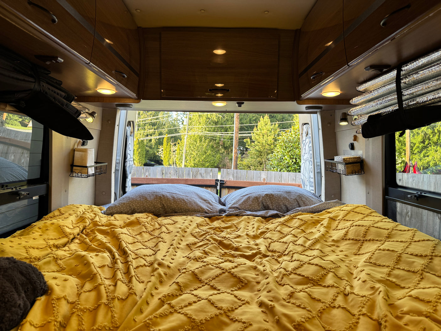 This RV rental features a cozy bed with a yellow textured comforter & gray pillows, offering a scenic view out the back.