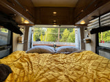 This RV rental features a cozy bed with a yellow textured comforter & gray pillows, offering a scenic view out the back.