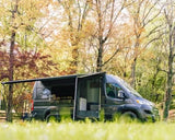 Modern dark grey camper van RV rental with an extended awning, parked in a sunny natural setting among trees.