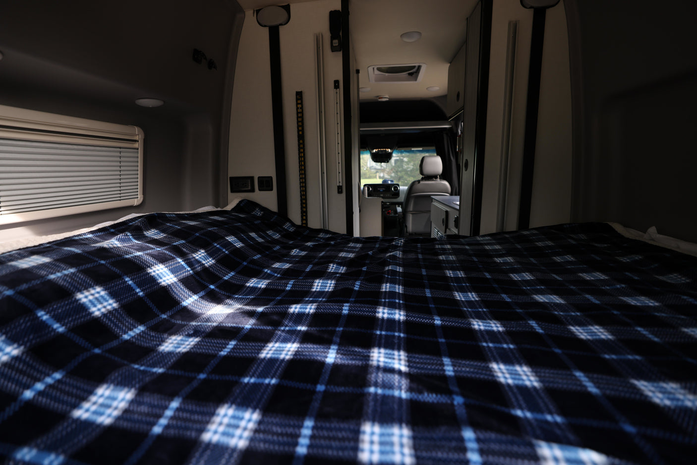 Inside an RV rental: a cozy bed with blue plaid blanket, looking forward to the driver's area. Ready for adventure.