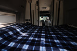 Inside an RV rental: a cozy bed with blue plaid blanket, looking forward to the driver's area. Ready for adventure.