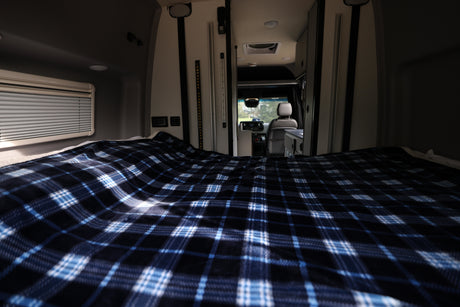 Inside an RV rental: a cozy bed with blue plaid blanket, looking forward to the driver's area. Ready for adventure.