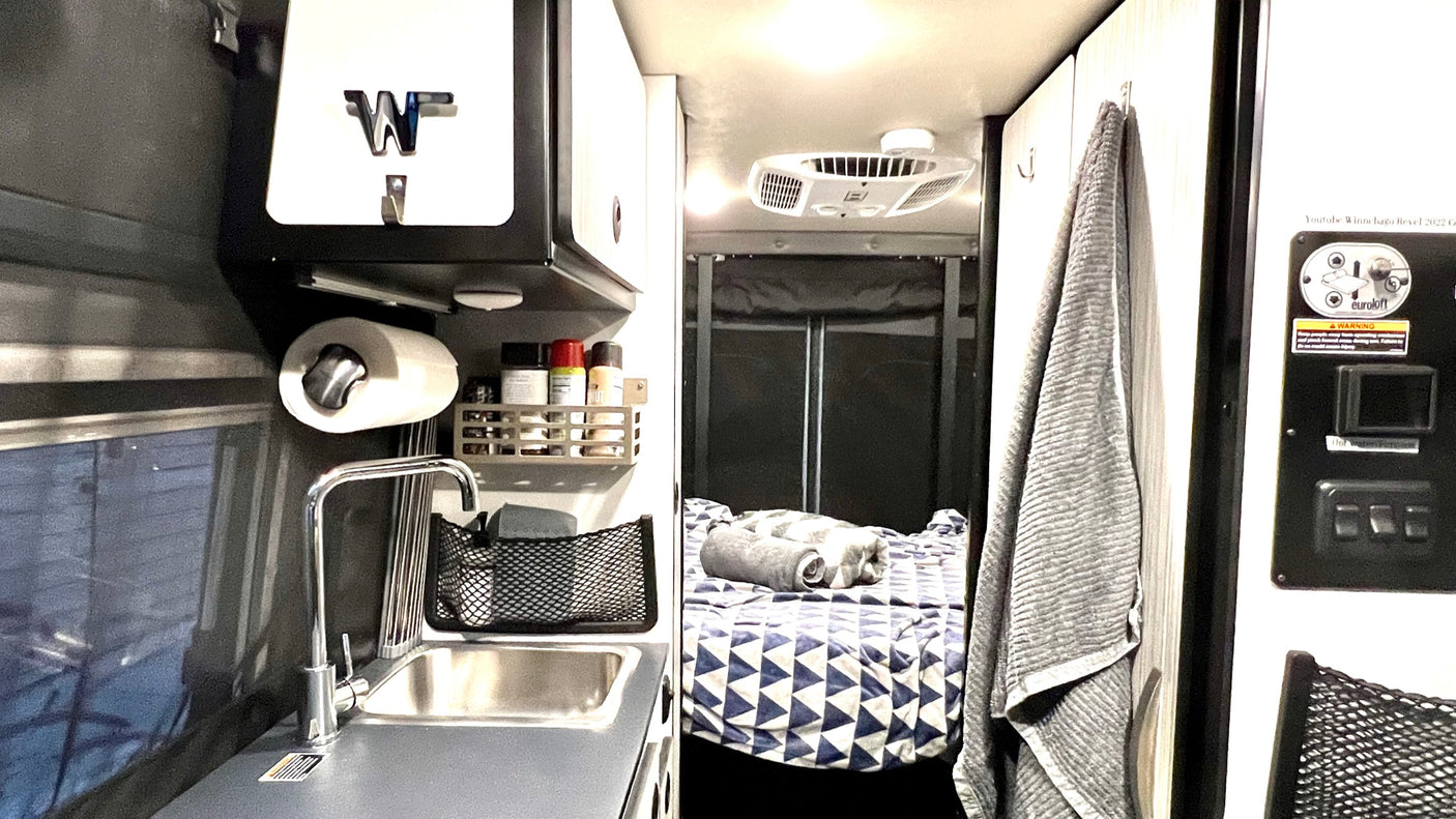 Cozy RV rental interior shows a compact kitchen with sink, paper towel holder, spices, and a geometric-patterned bed.