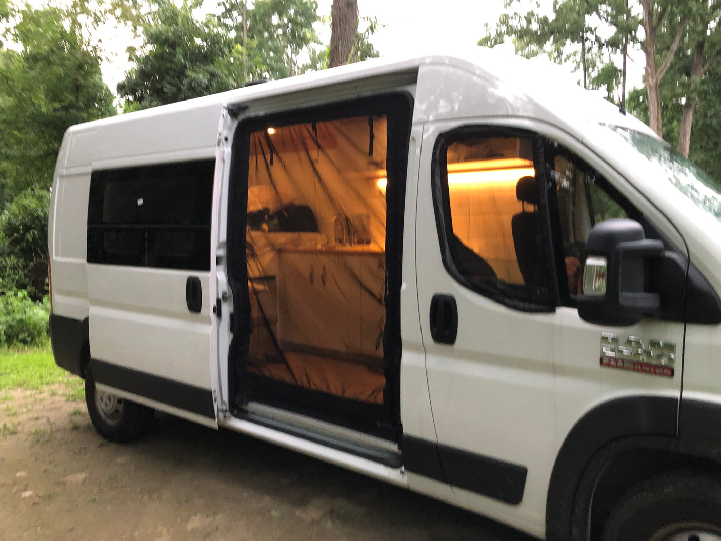 White **RV rental** camper van, side door open with screen, shows illuminated kitchen inside, parked outdoors among trees.