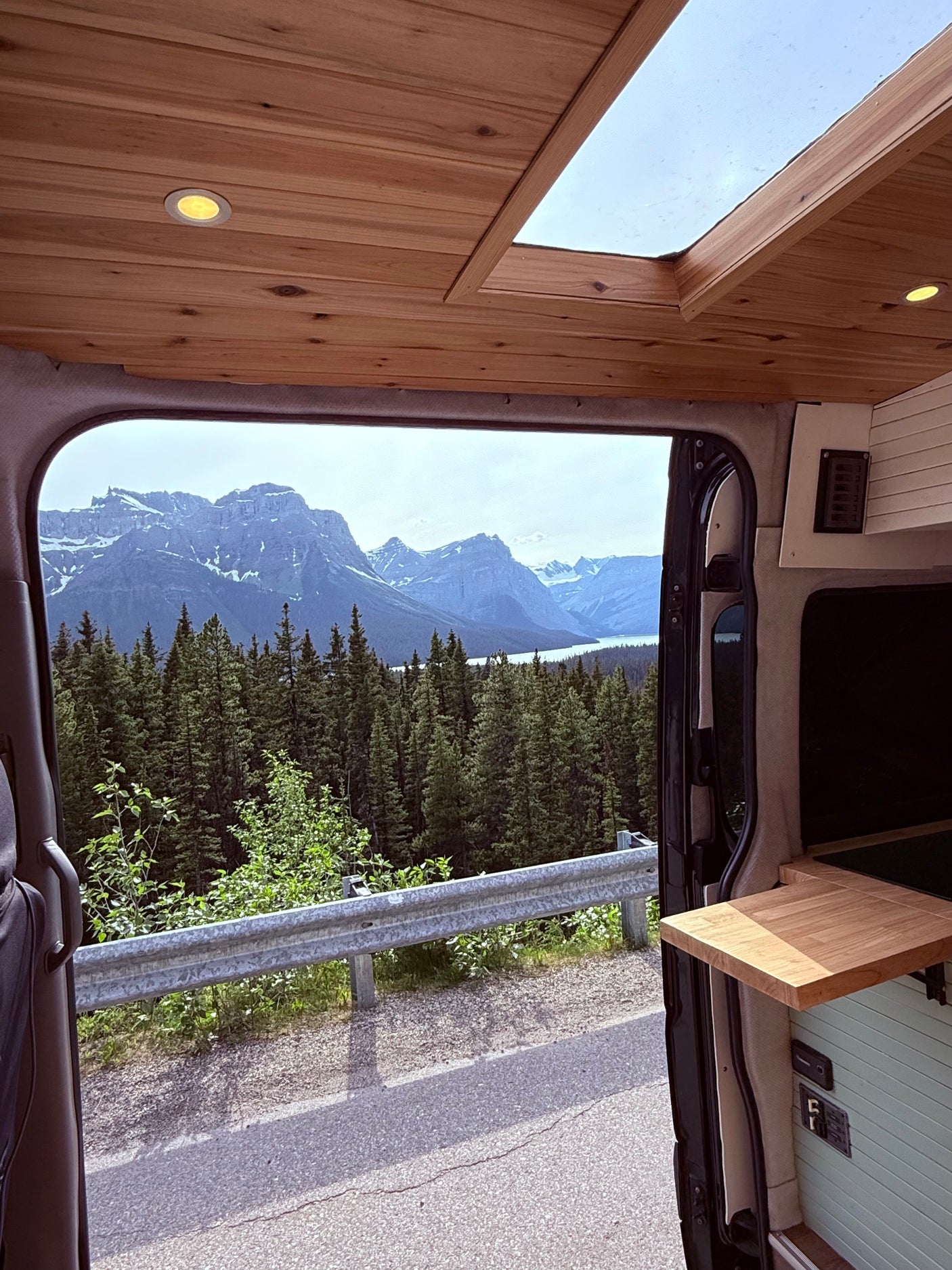 A cozy RV rental interior with wooden ceiling and skylight, offering a panoramic view of mountains, forests, and a lake.