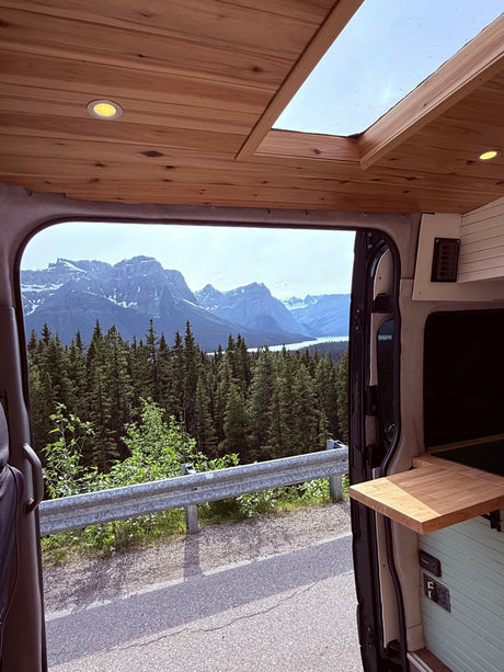 A cozy RV rental interior with wooden ceiling and skylight, offering a panoramic view of mountains, forests, and a lake.