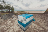 A blue and grey Dometic cooler rests on a sandy beach with rocks, ocean, and trees, ideal for your rv rental adventure.