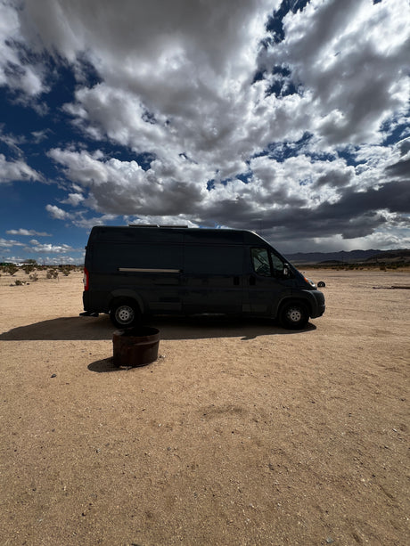 A dark RV rental van parked on sandy desert ground next to a fire pit under a dramatic cloudy sky. Ready for adventure!