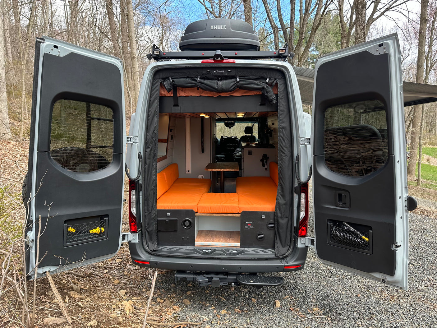 This custom camper van, ideal for rv rental, features an open rear with orange seating, a table, and a Thule roof box.