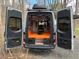 This custom camper van, ideal for rv rental, features an open rear with orange seating, a table, and a Thule roof box.