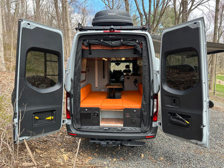 This custom camper van, ideal for rv rental, features an open rear with orange seating, a table, and a Thule roof box.