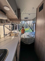 Inside a bright RV rental, a child on the bed gazes at a forest view, with a modern kitchen in the foreground.