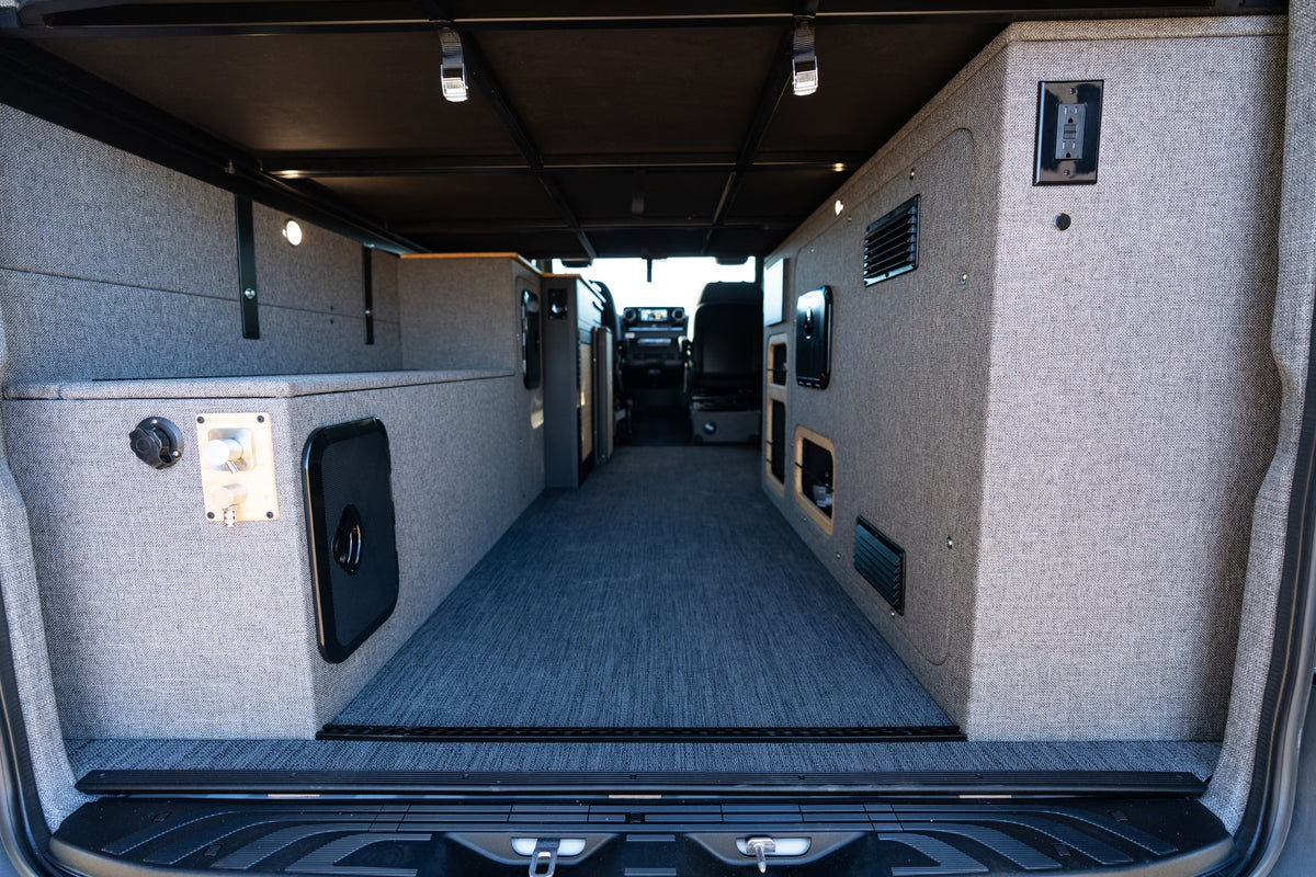 Custom RV rental interior features gray upholstered walls, integrated storage, carpet, and lighting, viewed from rear. — photo 13