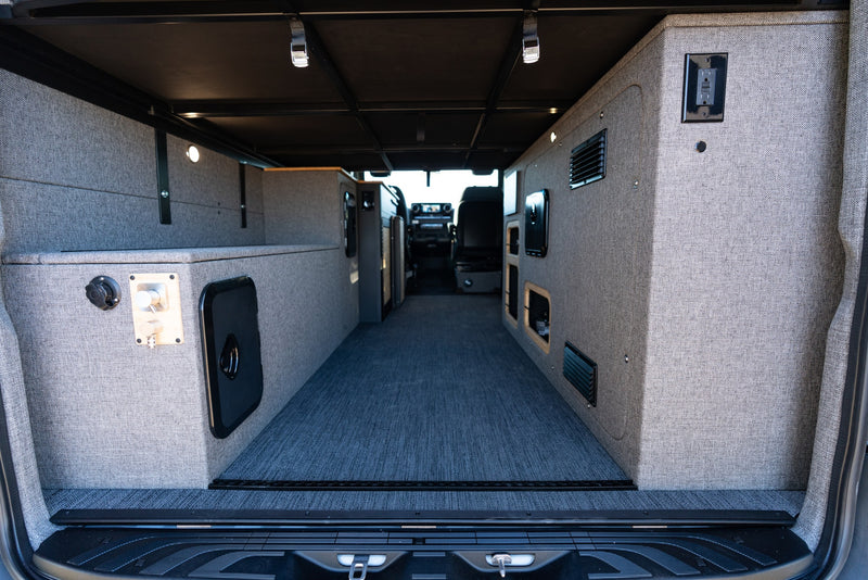 Custom RV rental interior features gray upholstered walls, integrated storage, carpet, and lighting, viewed from rear. — photo 13