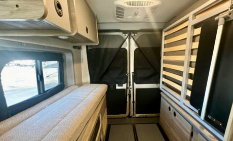 Interior of an rv rental: Left side bed, window, overhead storage. Rear doors with screens & a convertible bed frame.