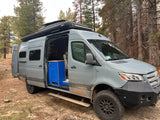 Custom off-road rv rental camper van in a forest, side door open to reveal blue interior cabinet & mud-splashed exterior.