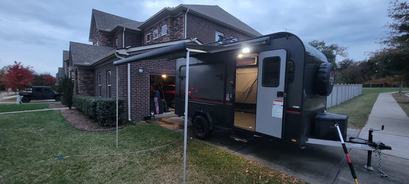 A dark RV rental travel trailer with an extended awning is parked in a residential driveway next to a brick house.