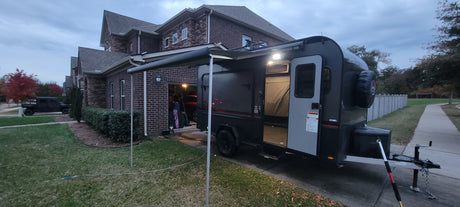 A dark RV rental travel trailer with an extended awning is parked in a residential driveway next to a brick house.