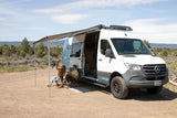 A family enjoys their RV rental parked in a scenic natural setting. The white Mercedes camper van features an awning, roof rack, and side step.