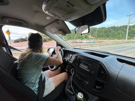Inside an RV rental, a person drives on a scenic coastal road with red cliffs and boats by the shore.