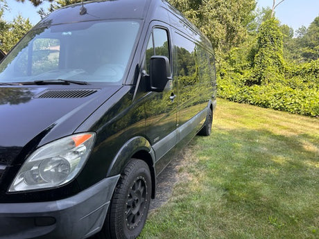 This black Sprinter-style van, perfect RV rental, is parked on a sunlit grassy area near dense foliage.