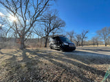 Sunny day campsite: Black camper van with extended awning under bare trees. Your perfect RV rental for outdoor adventures awaits!
