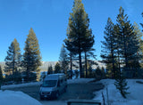 RV rental parked in a scenic winter landscape with a lake, snow-capped mountains, and tall pine trees under a blue sky.