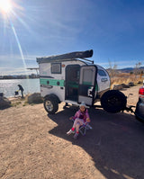A young girl enjoys the outdoors by an rv rental, a Sunray Sport camper, parked by a sunny lake. A person fishes nearby.