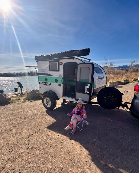A young girl enjoys the outdoors by an rv rental, a Sunray Sport camper, parked by a sunny lake. A person fishes nearby.
