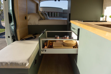 Modern RV rental interior: an open kitchen drawer displays dishes, utensils, and cutting boards; a cozy bed is visible.