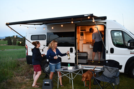 Friends gather around their RV rental camper van with an illuminated awning, enjoying an evening outdoors with drinks and a dog.