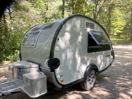 Silver teardrop **rv rental** camper with a unique rounded shape, parked on a dirt path under dappled sunlight in a forest. Ideal for camping.