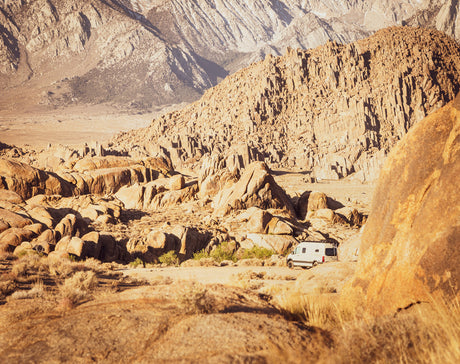An RV rental van is perfectly parked amidst large desert rock formations, with towering mountains in the distance.