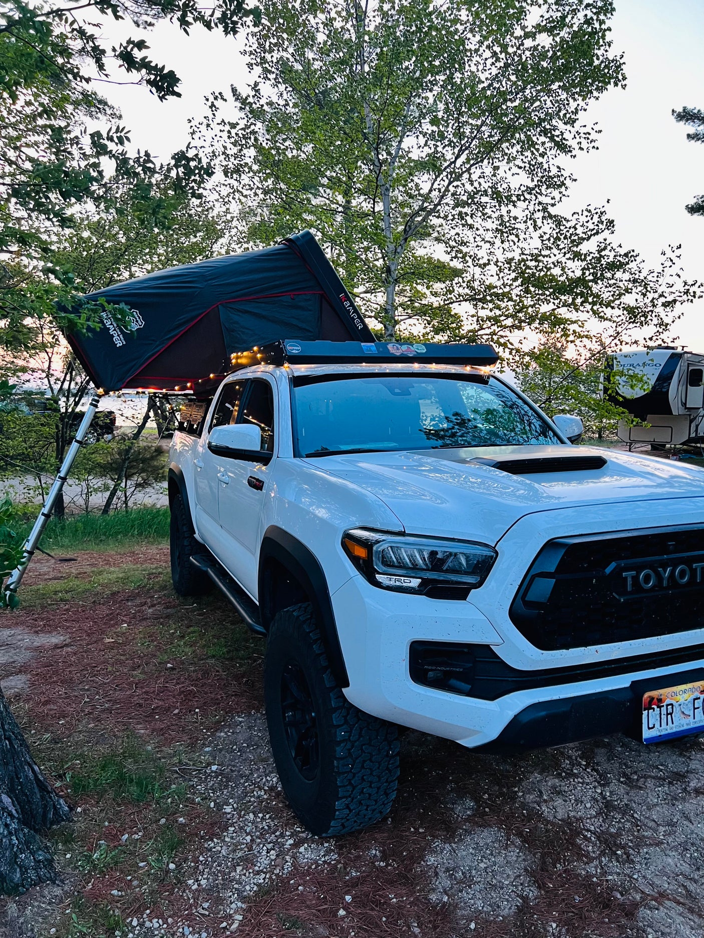 White Toyota Tacoma truck with an iKamper roof-top tent, set up for camping next to an RV rental by the water, for an overlanding adventure.
