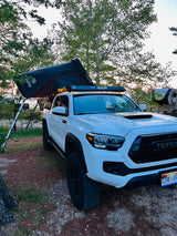 White Toyota Tacoma truck with an iKamper roof-top tent, set up for camping next to an RV rental by the water, for an overlanding adventure.