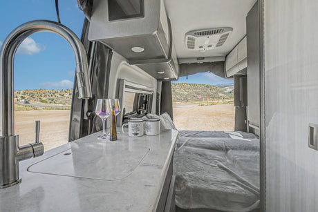 Modern RV rental interior featuring a kitchen with sink, wine glasses, mugs, a bed, and scenic desert views out the back.