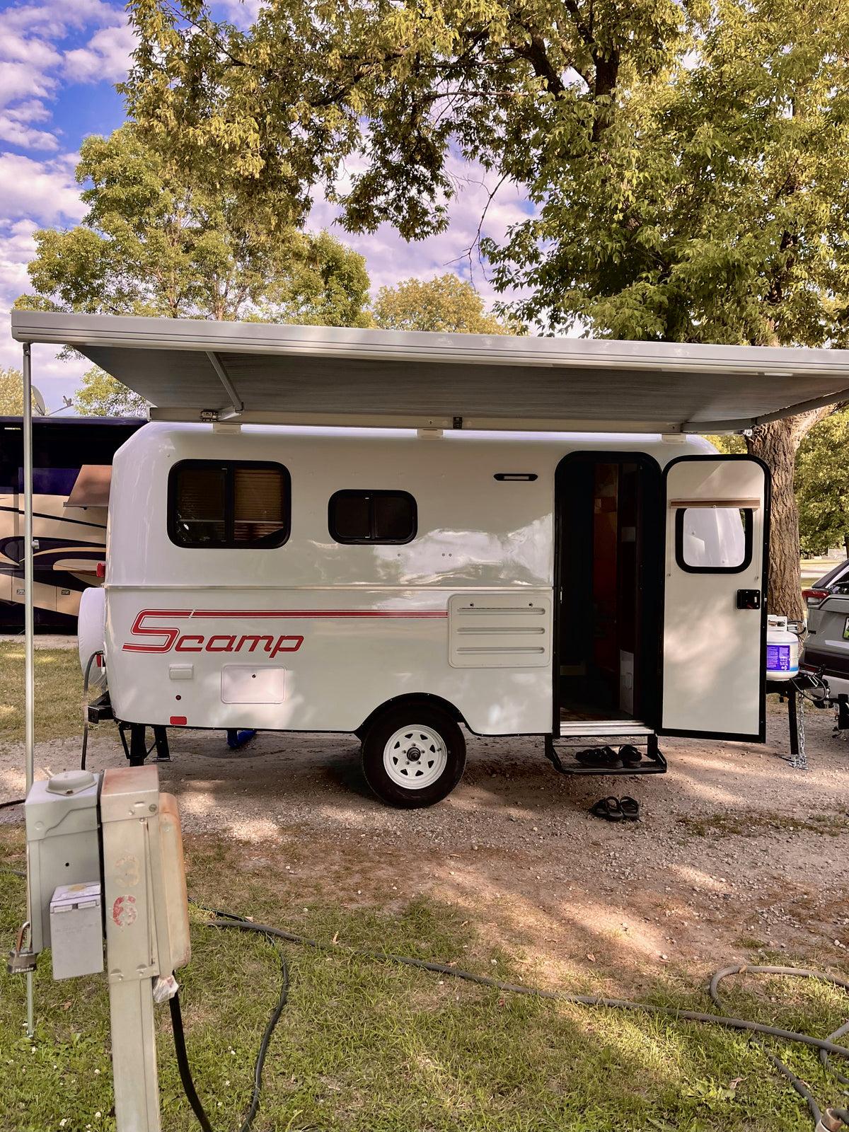 A white Scamp travel trailer with an extended awning at a leafy campground, perfect for an RV rental getaway. — photo 2