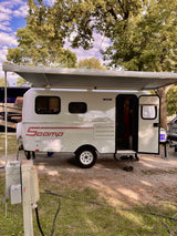 A white Scamp travel trailer with an extended awning at a leafy campground, perfect for an RV rental getaway.