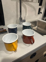 Four vibrant enamel camping mugs (yellow, navy, green, rust) on an RV rental kitchenette counter with a sink and faucet.