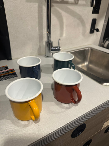 Four vibrant enamel camping mugs (yellow, navy, green, rust) on an RV rental kitchenette counter with a sink and faucet.