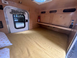 Interior view of a compact rv rental camper showing a cozy bed, side door with window, overhead storage, and a fire extinguisher.