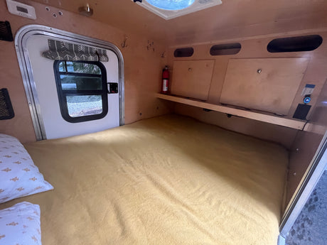 Interior view of a compact rv rental camper showing a cozy bed, side door with window, overhead storage, and a fire extinguisher.