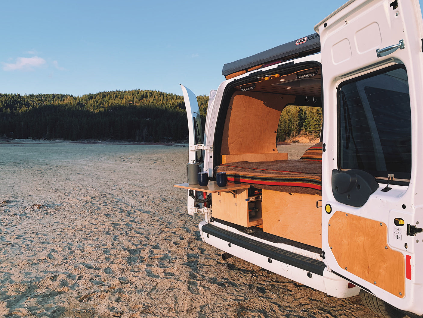 This customized camper van, ideal for an rv rental, is parked on a sandy shore with open doors revealing a bed and setup, facing a forest under a clear sky.