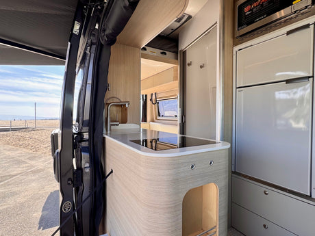 Modern RV rental interior showing a fully equipped camper van kitchen and open door to a beautiful beach view.