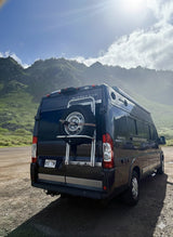 Black Winnebago RV rental with a spare tire, parked on a dirt road before scenic green mountains under a bright sky.