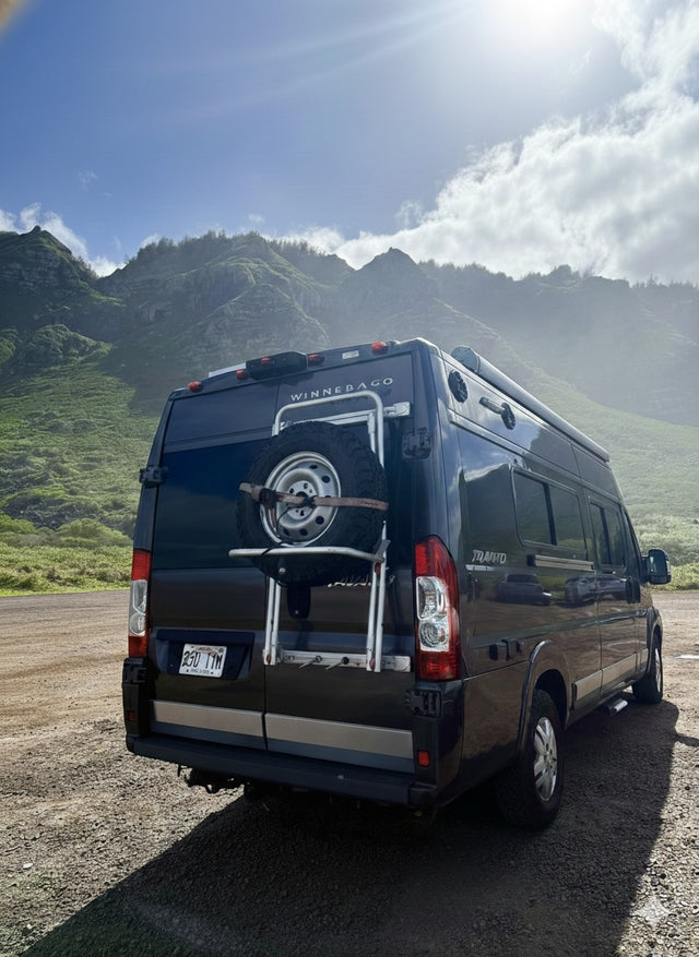 Black Winnebago RV rental with a spare tire, parked on a dirt road before scenic green mountains under a bright sky.